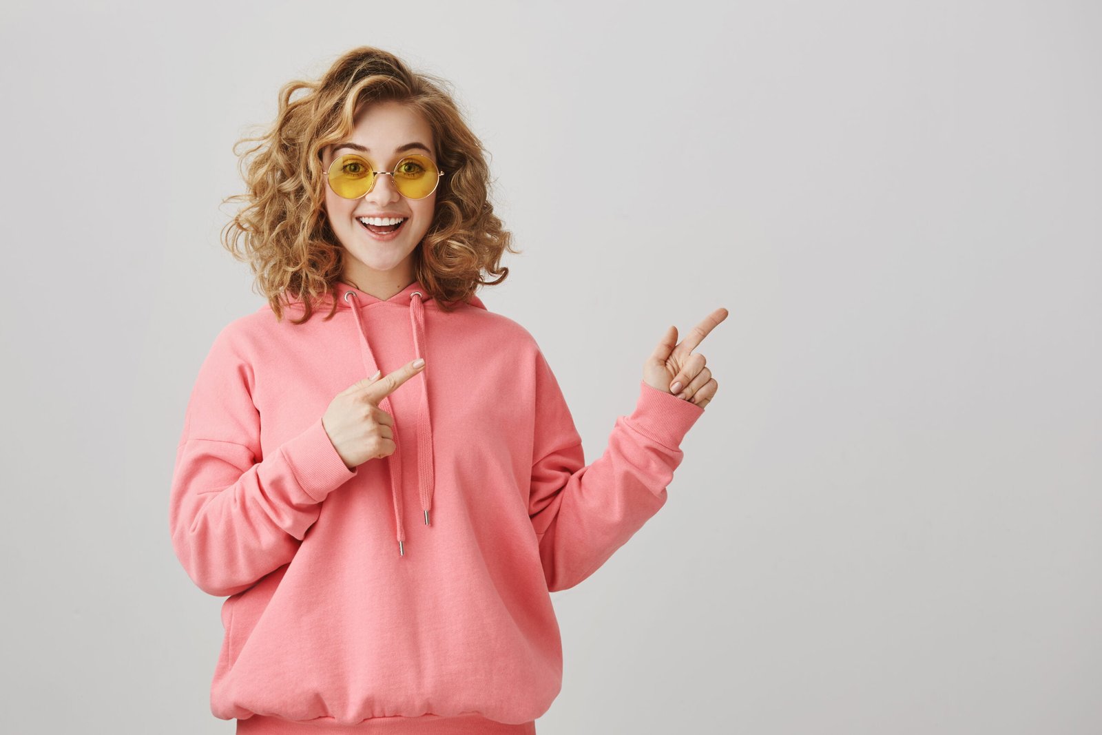 About Us 6 Shall we hit this bar and dance all night. Stylish attractive young woman with short curly hairstyle and trendy sunglasses pointing right with index fingers and smiling with interest over gray wall.